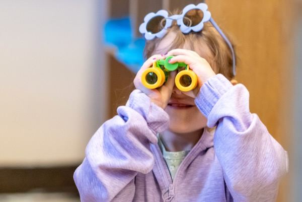 Young girl wearing a lavender sweater looking through toy binoculars.