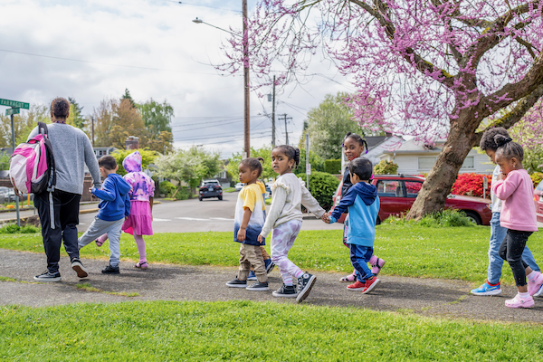 Several children walking in an orderly line while holding hands as they follow their teacher through a park..