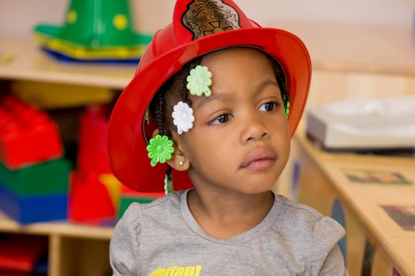 Young girl wearing a red toy firefighter's helmet.