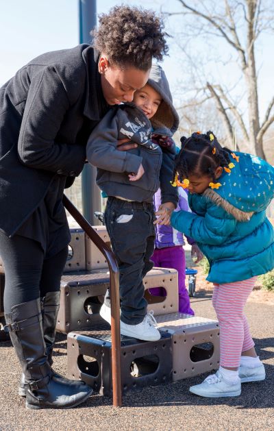 Young child being helped down playground stairs by teacher and another classmate.